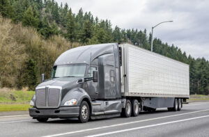 Stylish gray big rig semi truck tractor transporting cargo in refrigerator semi trailer running on the interstate highway road with forest line on the side
