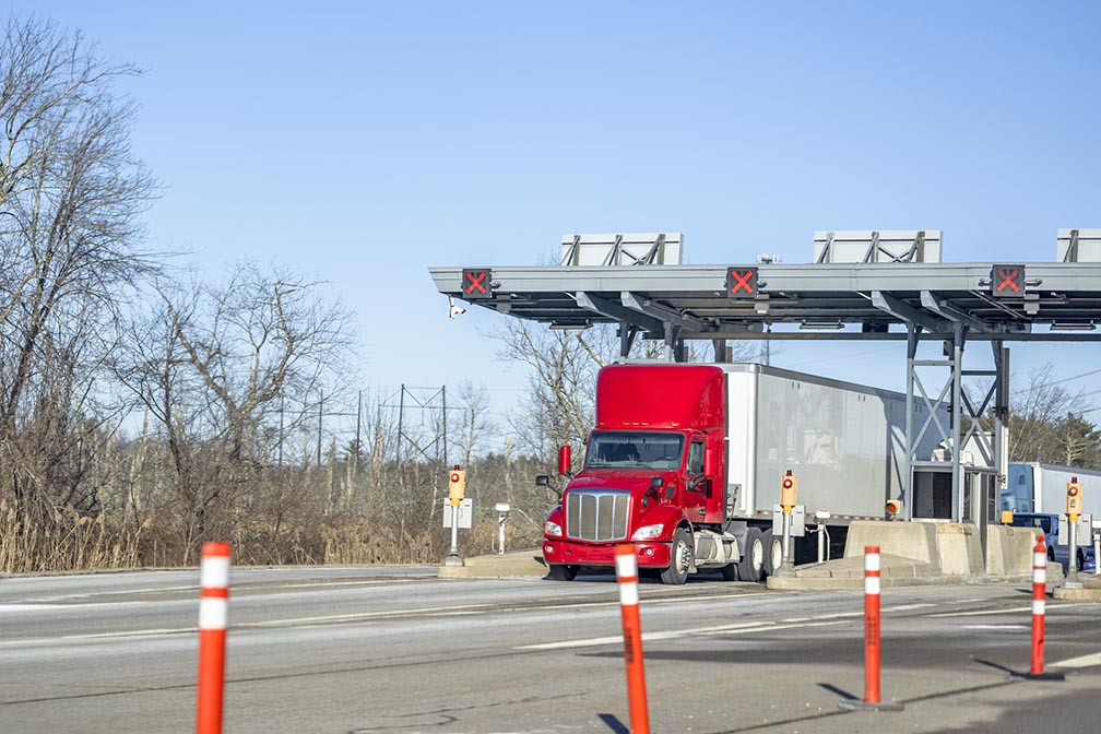 Red day cab big rig semi truck with dry van semi trailer passes a toll booth on a toll highway road