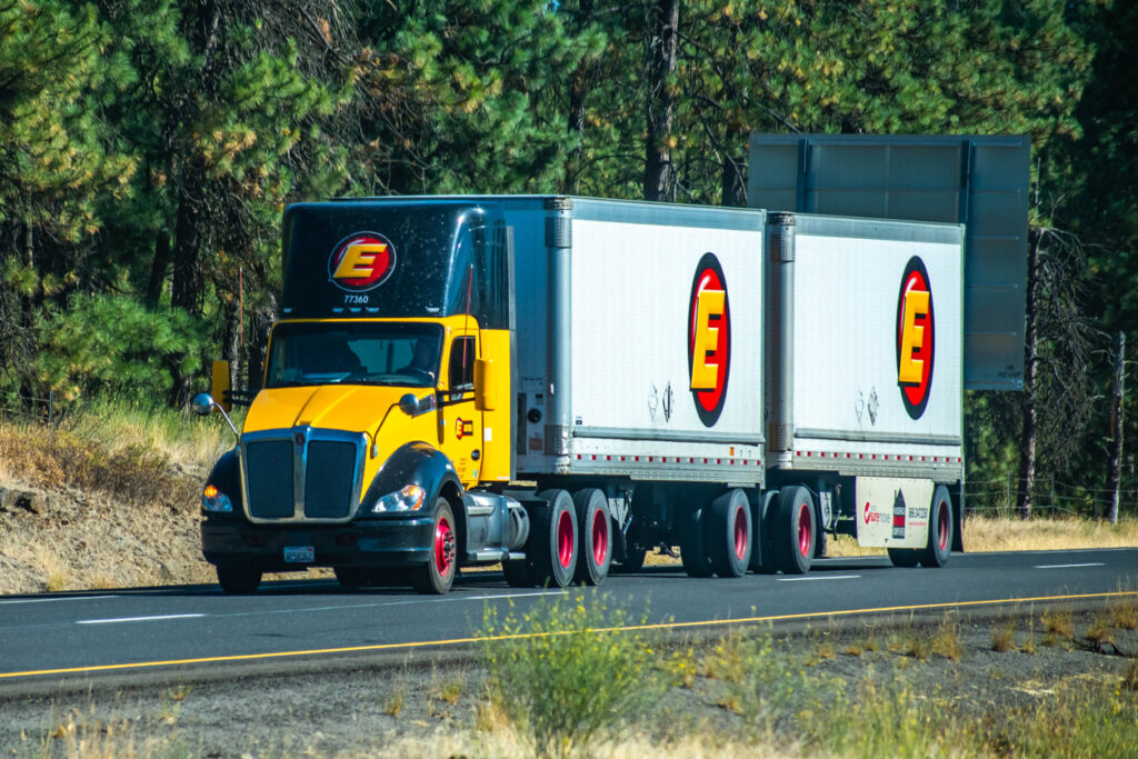 Estes Express Lines Semi Truck with double trailers driving on a wooded section of the Interstate, hauling freight in between metropolitan areas.