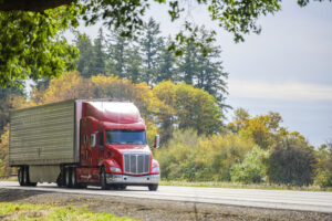 Bright big rig red semi truck in sunshine transporting frozen and chilled foods in refrigerator semi trailer running on the straight highway road with green trees on the side
