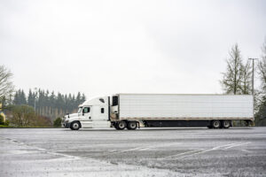 Professional bonnet white big rig semi truck with refrigerator semi trailer standing on the industrial marked parking lot at rainy weather with gray sky