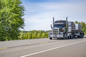 Classic black big rig semi truck with aluminum protective grille guard and chrome accessories transporting fastened lumber wood on flat bed semi trailer driving on the flat highway road